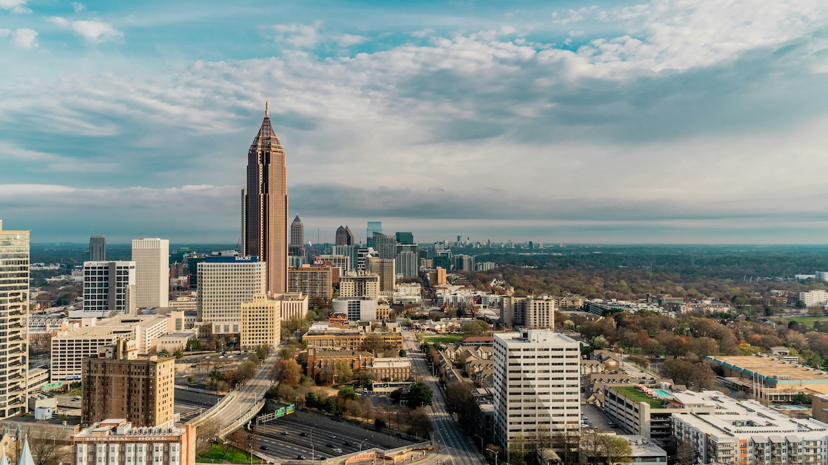 Aerial view of downtown Atlanta, Georgia, featuring tall skyscrapers including the Bank of America Plaza tower, surrounded by mid-rise buildings, highways, and tree-filled neighborhoods under a partly cloudy sky.