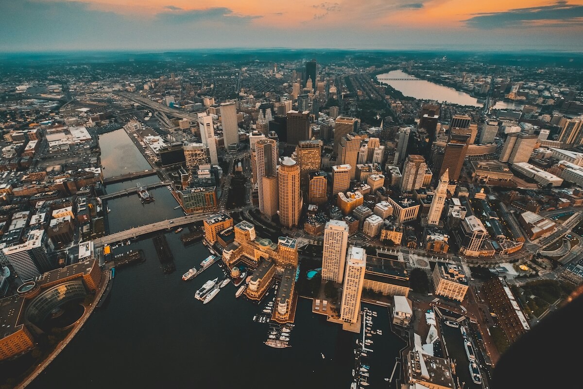Aerial view of Boston’s cityscape at sunset, showing skyscrapers, historic buildings, bridges, and boats docked along the waterfront with the Charles River in the background.