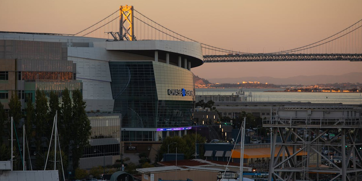 Exterior of Chase Center in San Francisco with the Bay Bridge in the background, captured at sunset. The arena’s curved glass facade reflects the warm evening light.