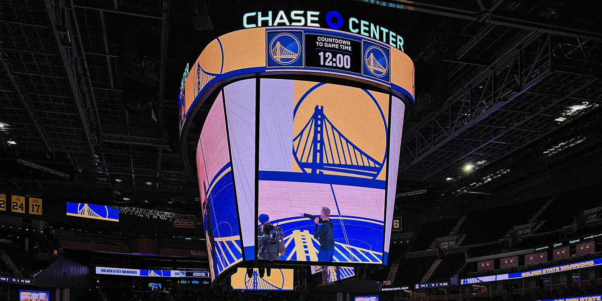 Interior view of Chase Center showing the giant center-hung scoreboard lit with Golden State Warriors logos and a countdown to game time. Two staff members stand below pointing at the display.