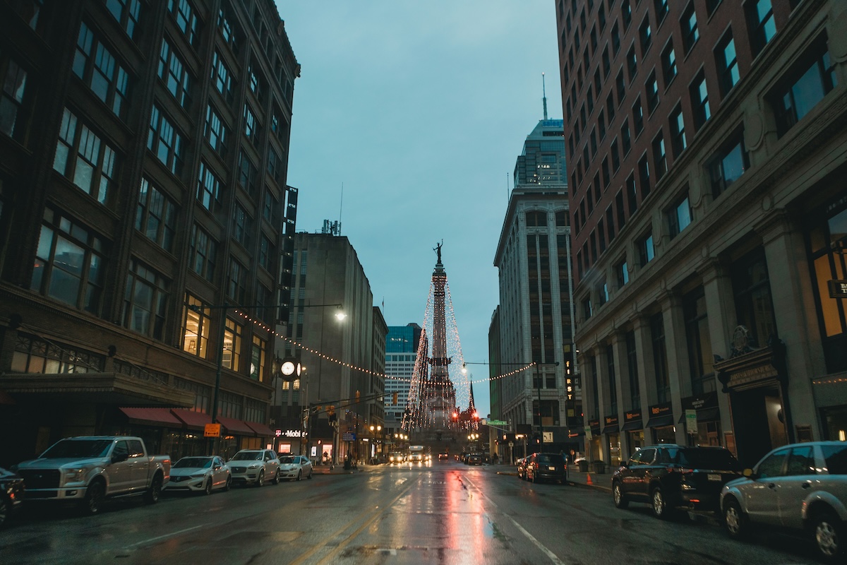 Downtown Indianapolis street view at dusk, with cars parked along wet pavement, tall buildings on both sides, and the Soldiers and Sailors Monument lit with holiday lights in the distance.