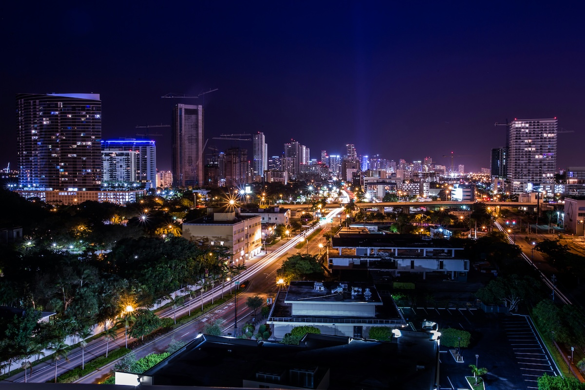 Miami skyline at night with brightly lit skyscrapers, illuminated streets, and light trails from traffic stretching into the distance.
