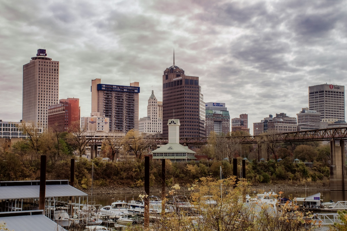 Downtown Memphis skyline with tall office buildings, trees with autumn foliage, and boats docked along the waterfront under a cloudy sky.