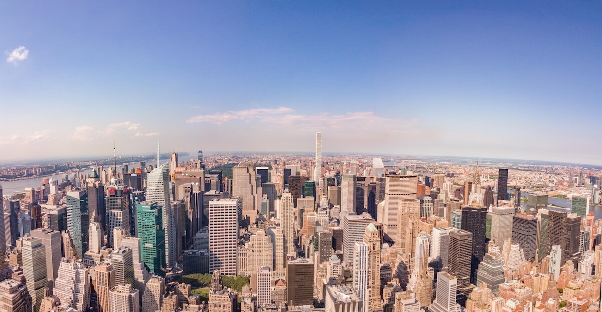 Aerial panoramic view of the New York City skyline on a clear day, showcasing a dense cluster of skyscrapers in Midtown Manhattan, with the Hudson River visible to the left and the East River to the right.