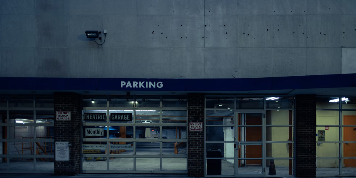 Exterior view of a parking garage entrance labeled 'PARKING,' with glass doors and signs indicating monthly parking and tow-away zones.