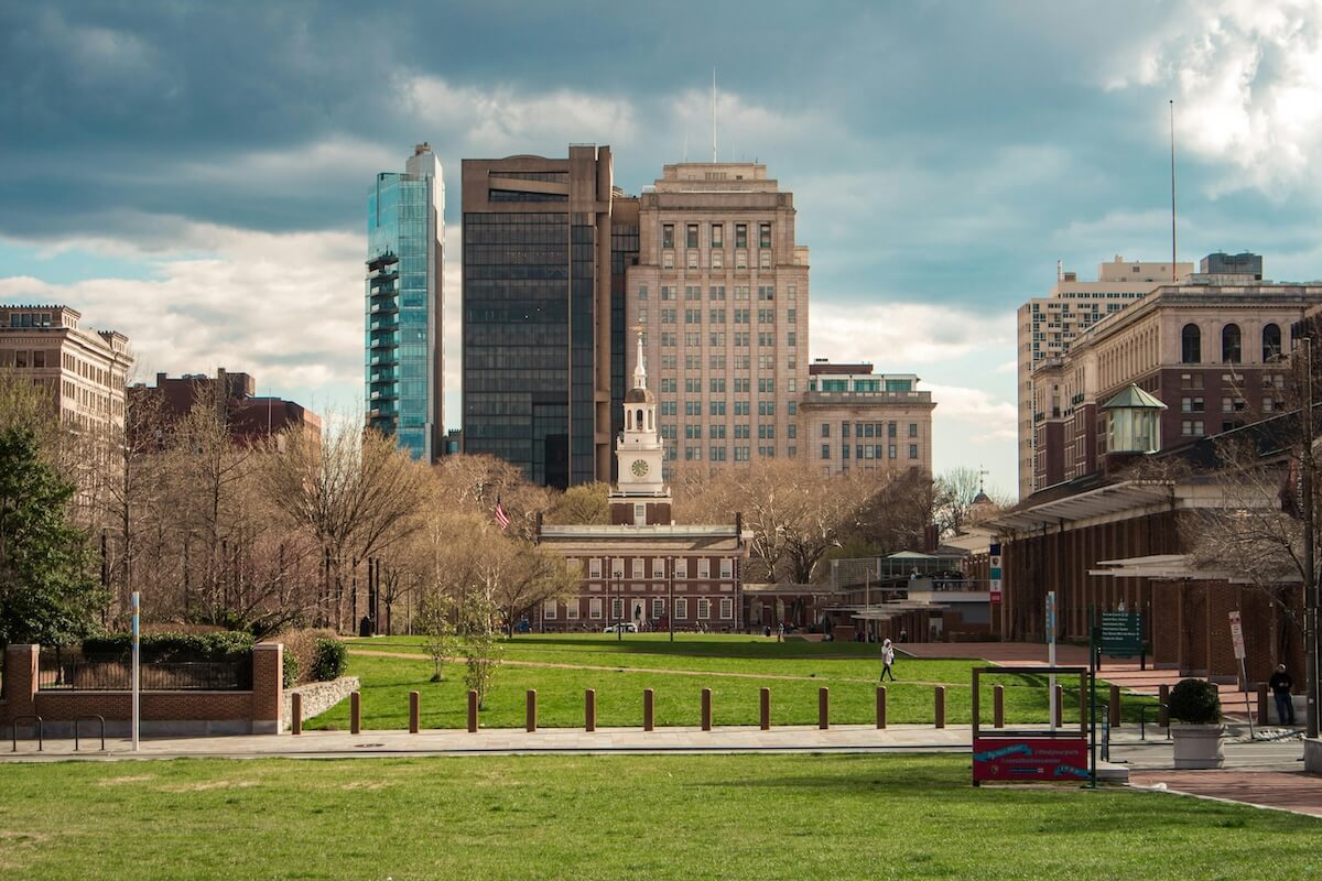 View of Independence Hall in Philadelphia with its iconic clock tower, surrounded by modern skyscrapers and trees on a partly cloudy day. A few people walk across the green lawn in the foreground.