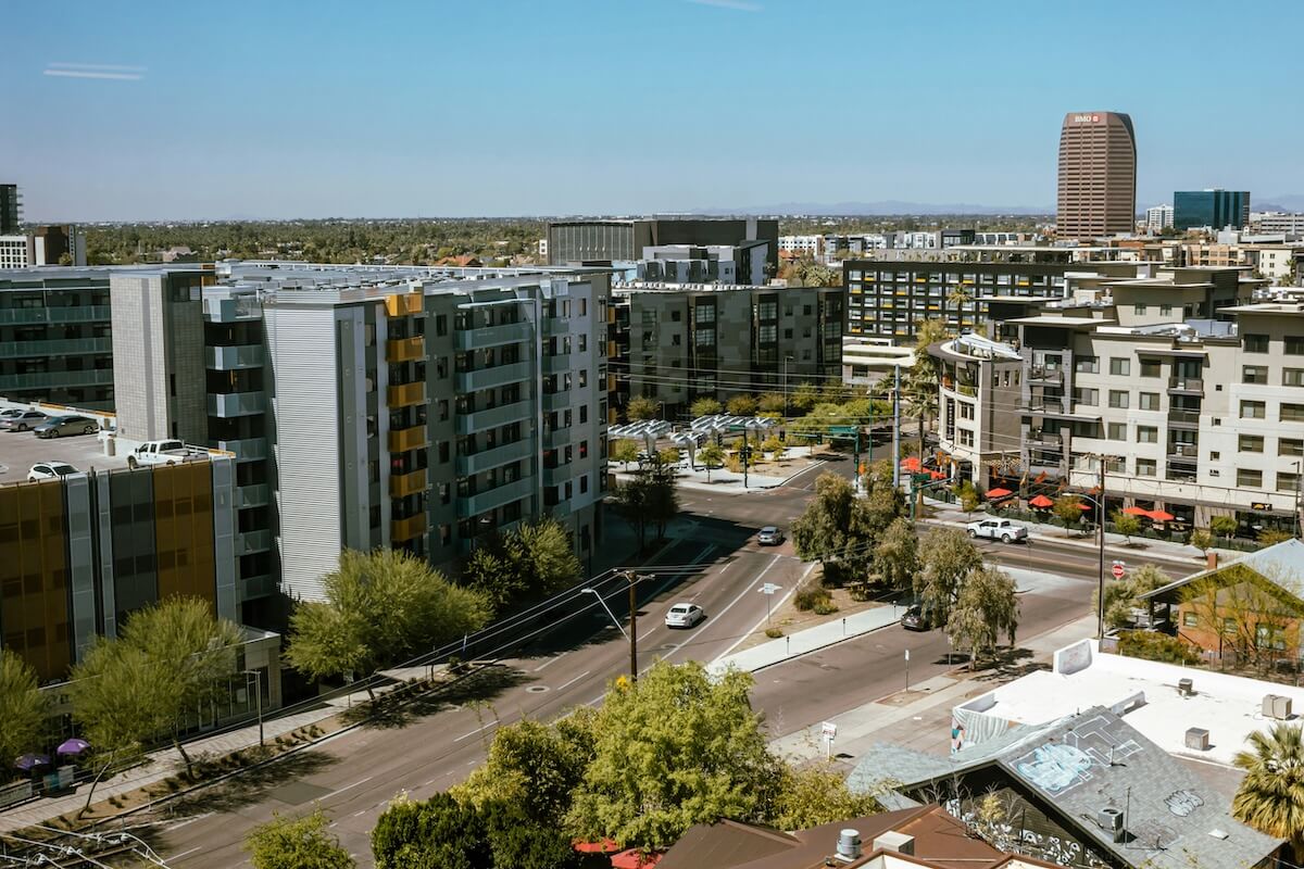 A daytime cityscape of Phoenix, Arizona, featuring modern mid-rise apartment buildings with balconies, a few taller office towers in the distance, and desert trees along the streets under a clear blue sky.