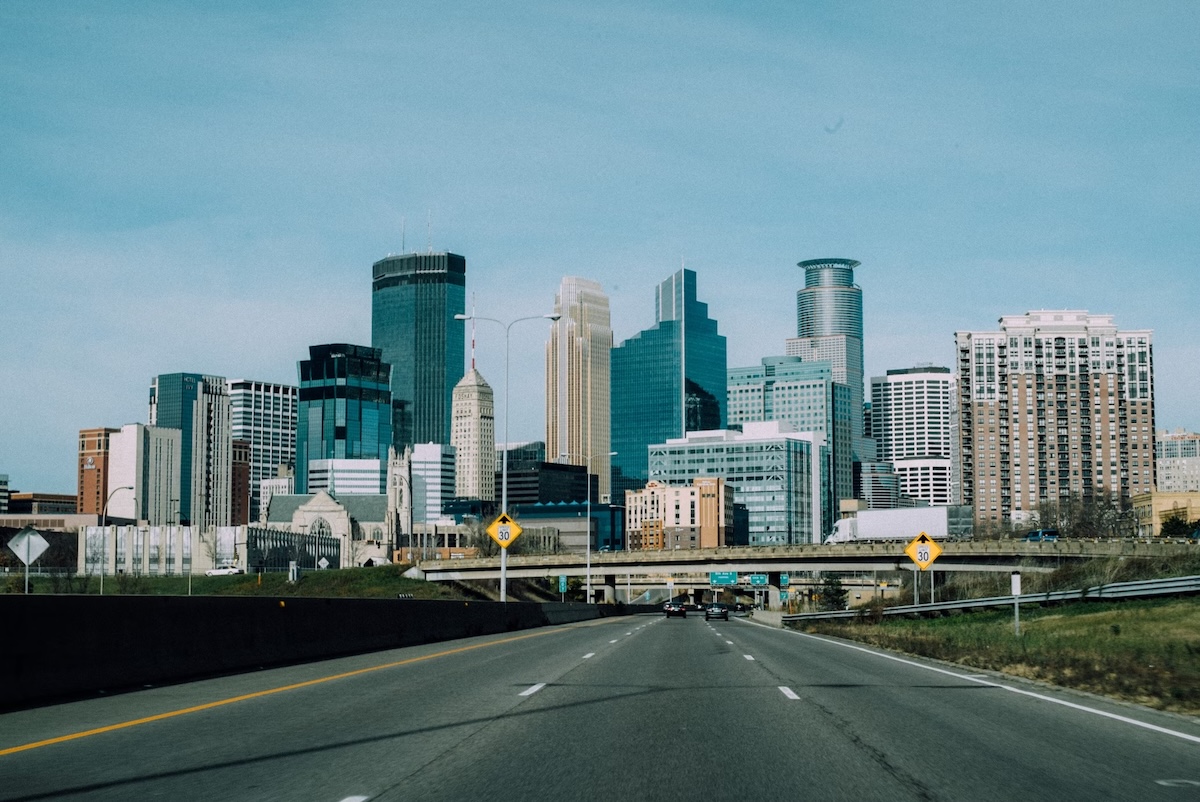 View of downtown Minneapolis, Minnesota, showing a cluster of modern skyscrapers and office buildings as seen from a highway leading into the city.