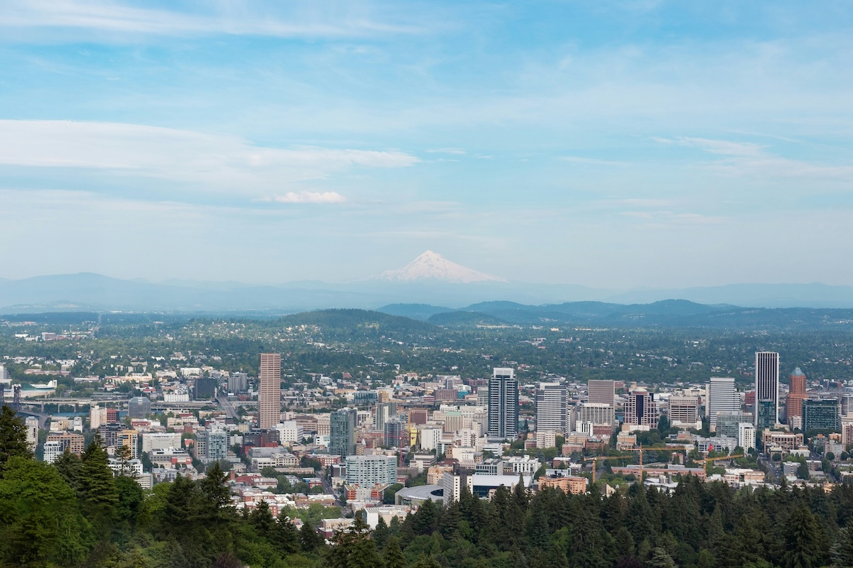 A panoramic view of downtown Portland, Oregon, with modern high-rise buildings surrounded by lush greenery, and Mount Hood’s snow-capped peak visible in the distance under a clear blue sky.