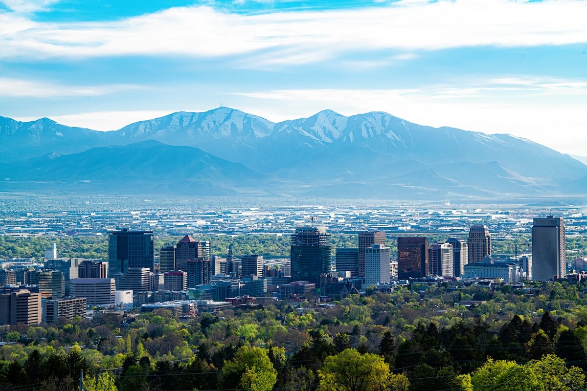 A scenic view of downtown Salt Lake City, Utah, with modern skyscrapers rising above a lush, green landscape, set against the backdrop of the snow-capped Wasatch Mountains under a bright blue sky.