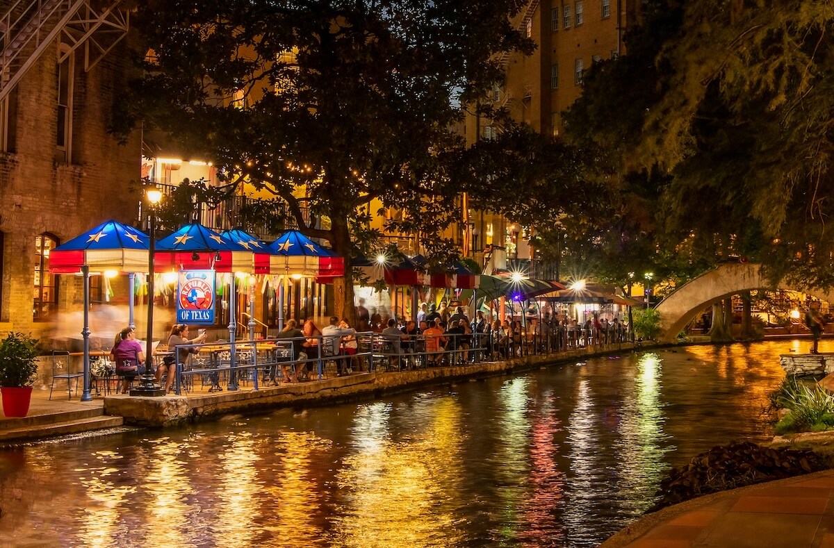 A lively nighttime scene along the San Antonio River Walk in Texas, with colorful umbrellas and string lights illuminating outdoor restaurant seating beside the water, where reflections shimmer under a stone arch bridge.