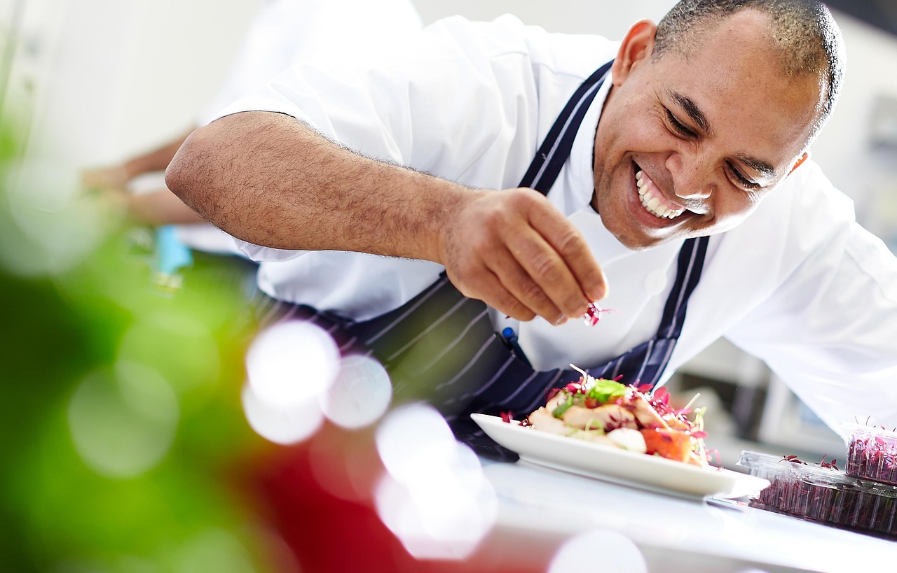 Smiling chef in a professional kitchen carefully garnishing a plated dish with microgreens, focused on presentation and fine dining details.