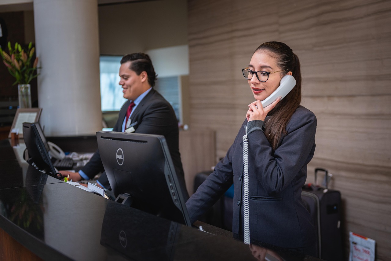 Hotel front desk with two reception staff assisting guests; a woman in glasses speaks on a corded phone while a colleague works at a computer behind the counter.